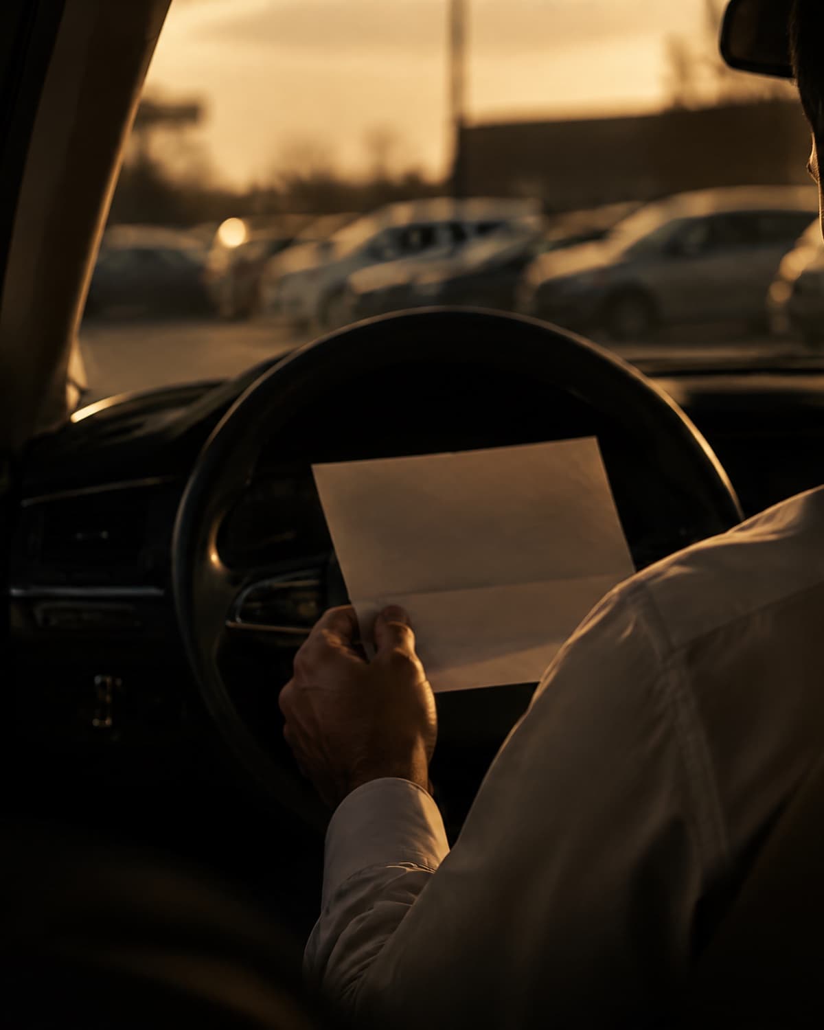 A man sitting in his parked car at dusk, holding a folded letter from his employer.