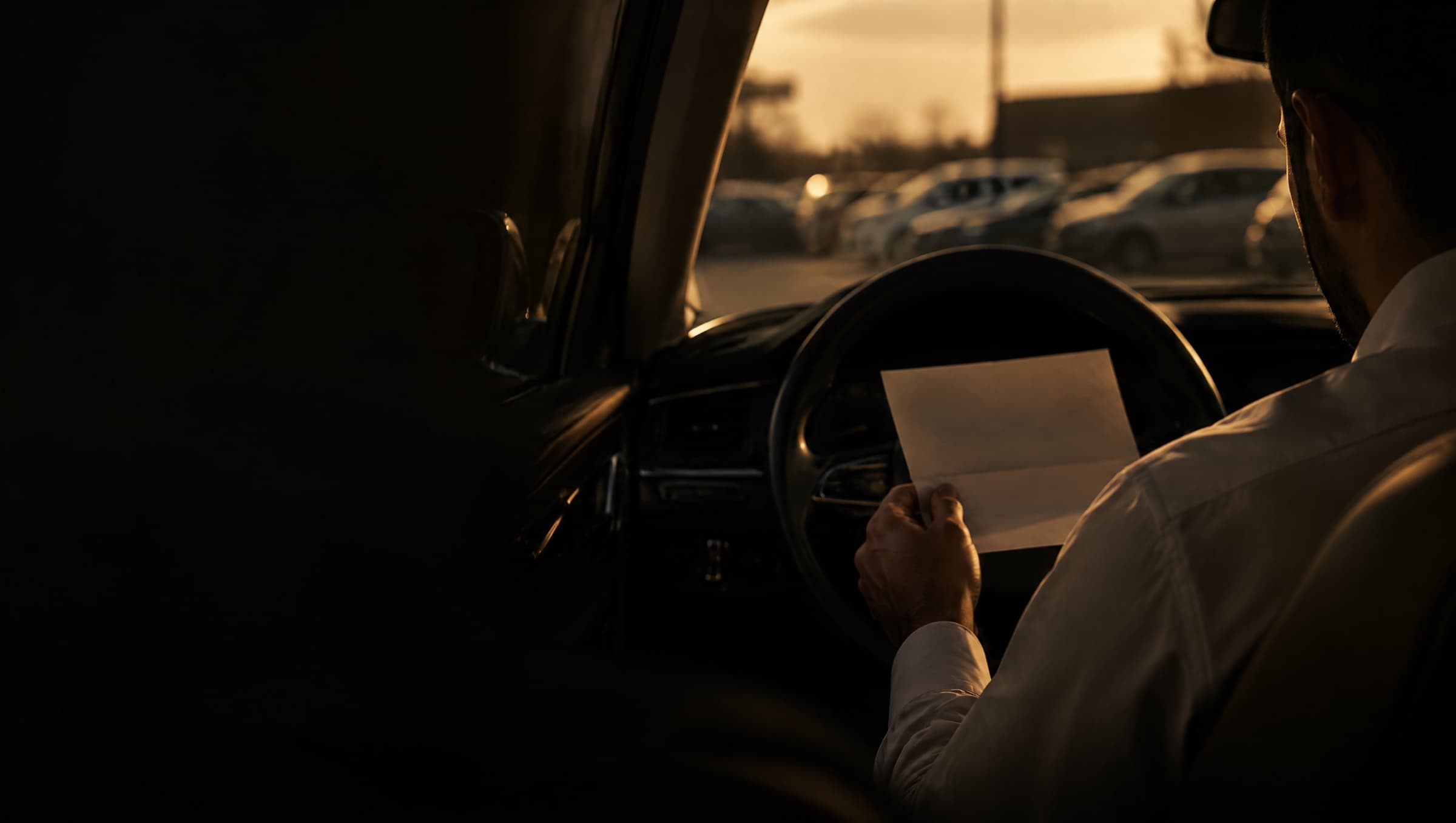 A man sitting in his parked car at dusk, holding a folded letter from his employer.